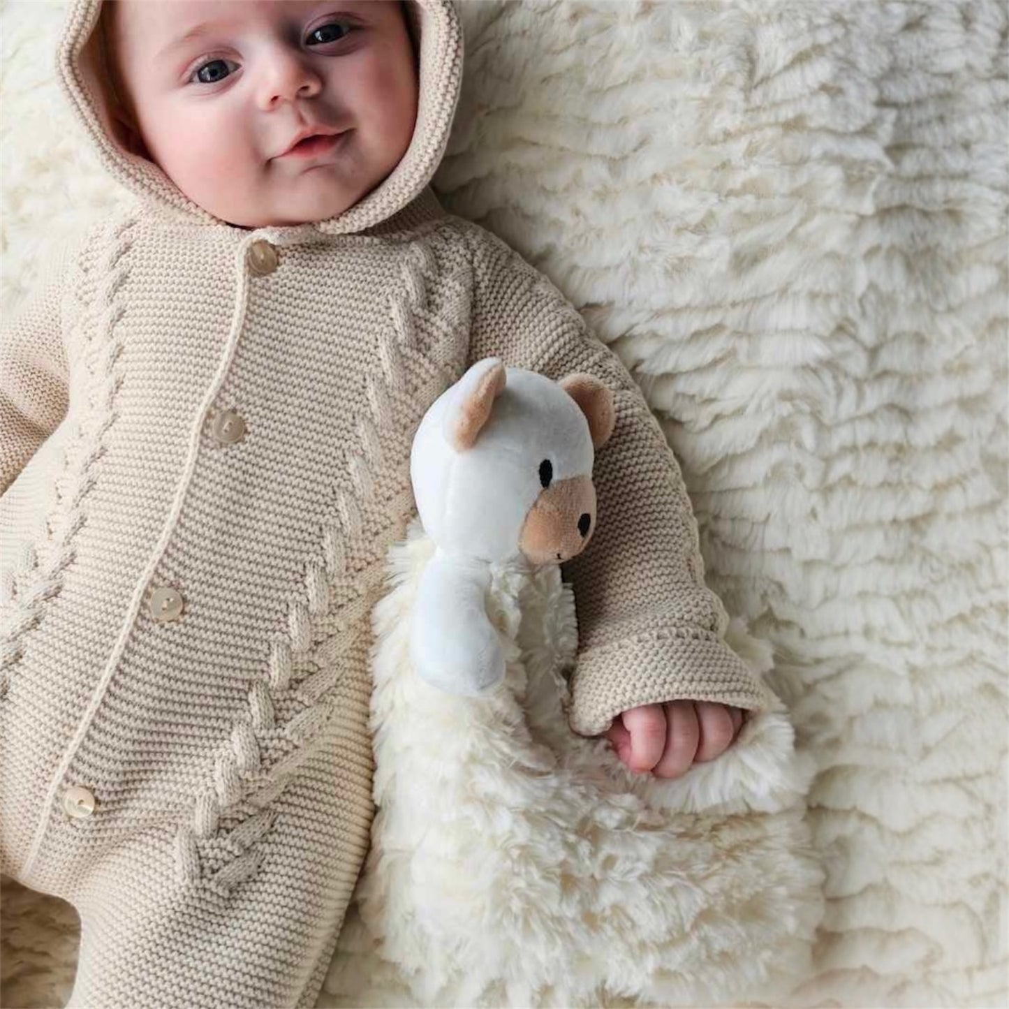 Baby lying on a soft cream teddy blanket holding a bear comforter while wearing a taupe knit romper