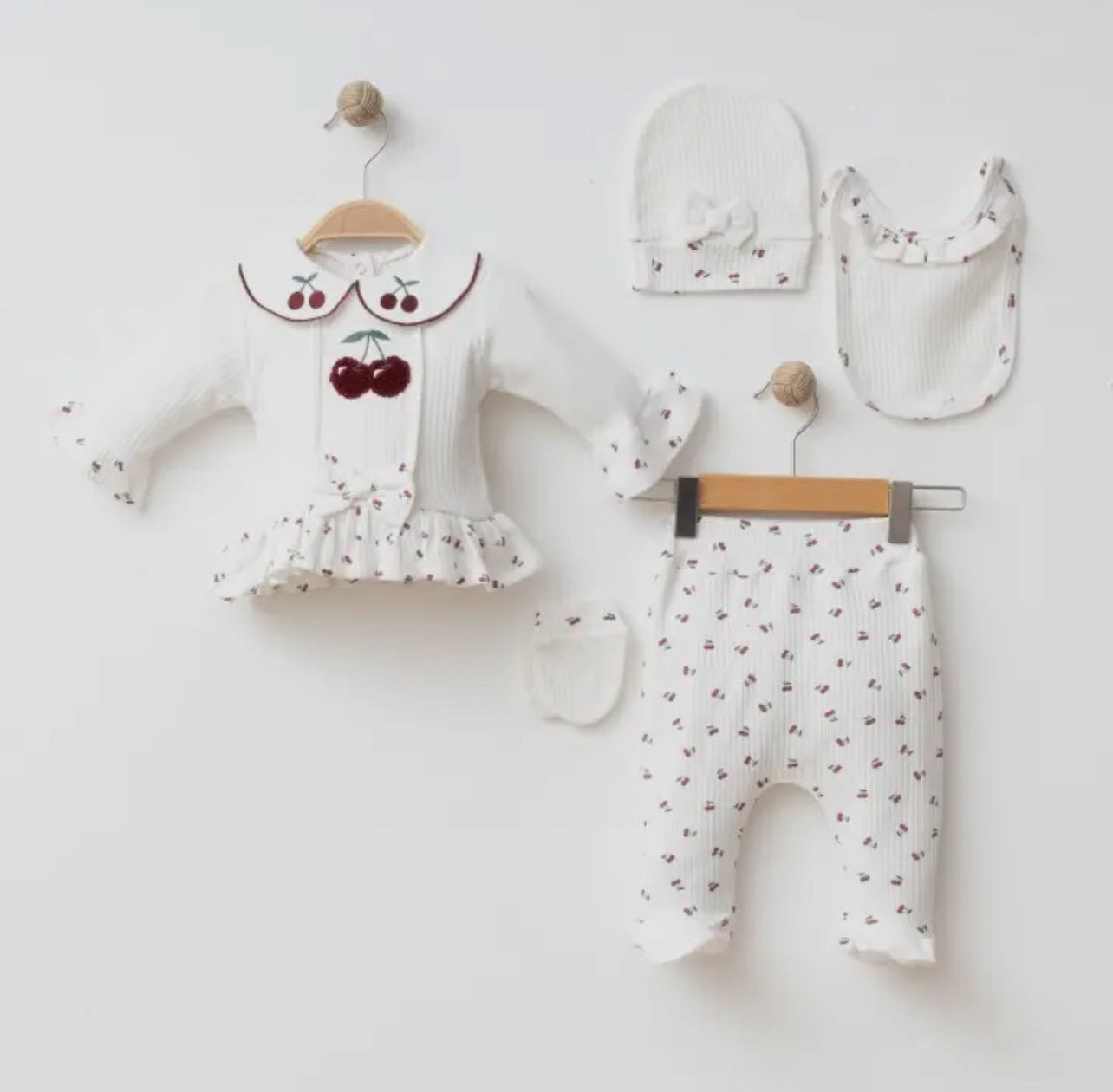 Flat lay of the 5-piece Cherry Blossom baby set in white with red cherry patterns, featuring a ruffled top, leggings, beanie, bib, and mitten.