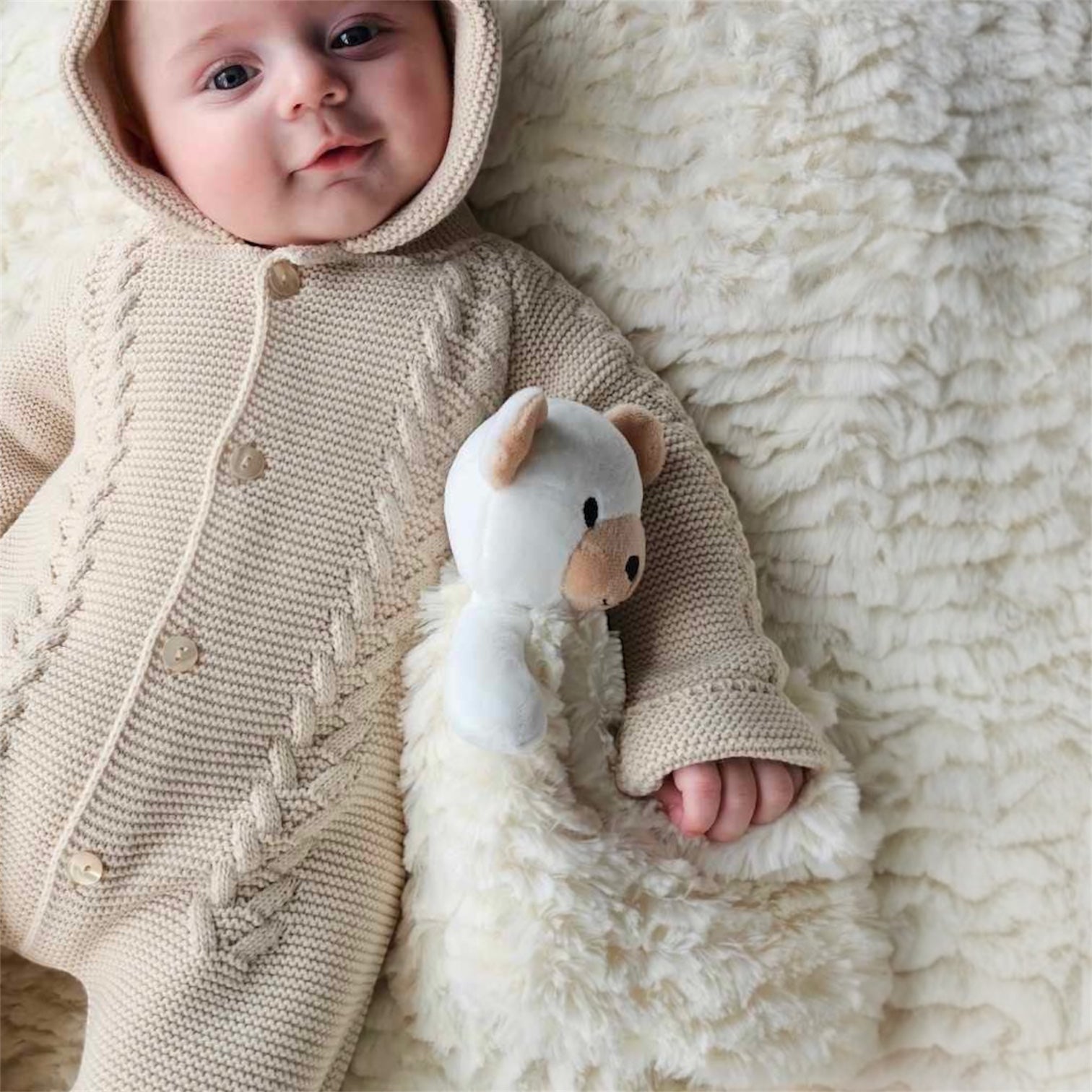 Baby lying on a soft cream teddy blanket holding a bear comforter while wearing a taupe knit romper