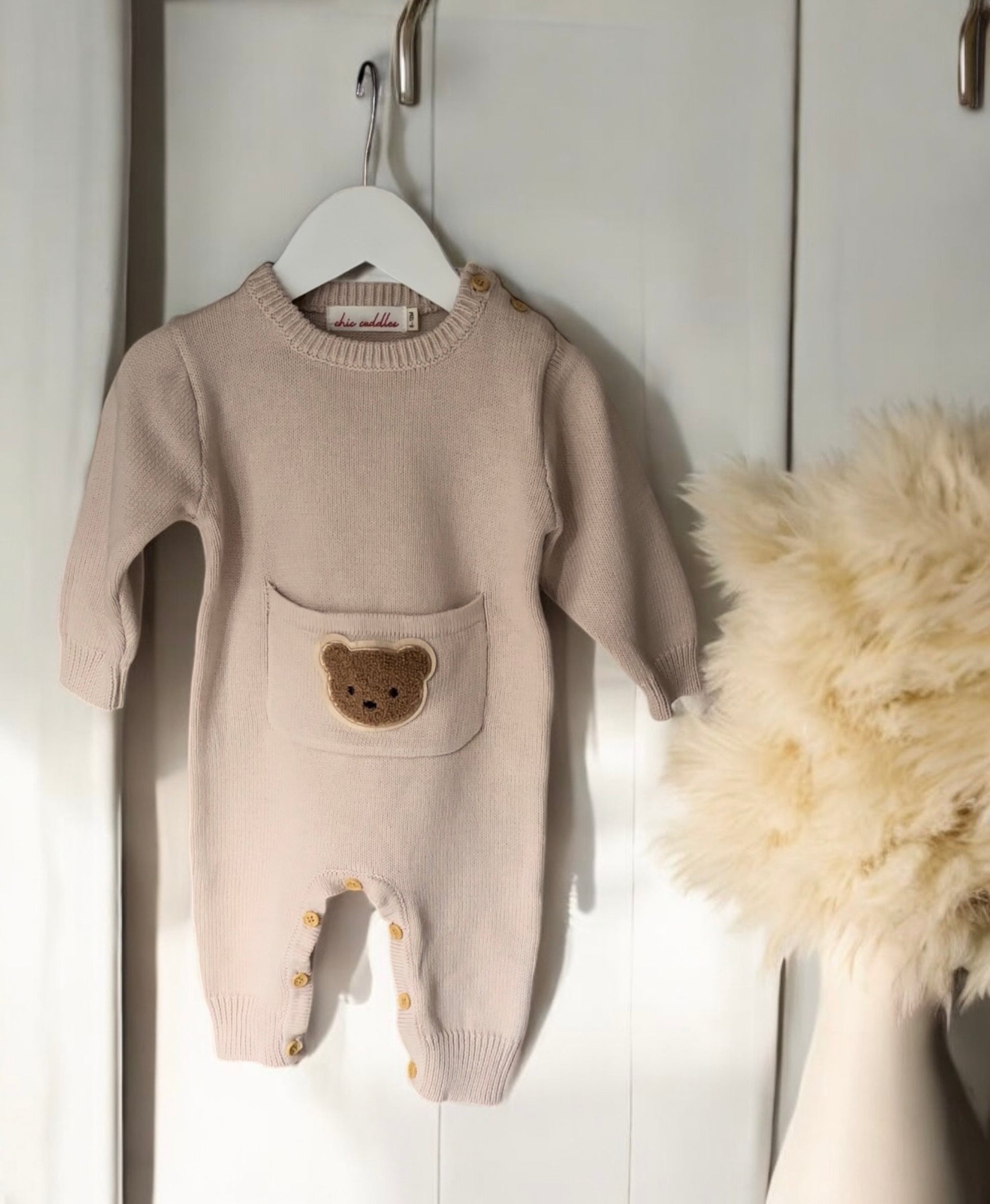Beige Cosi Bear Pocket Onesie displayed on a hanger, showing soft fabric and teddy bear pocket