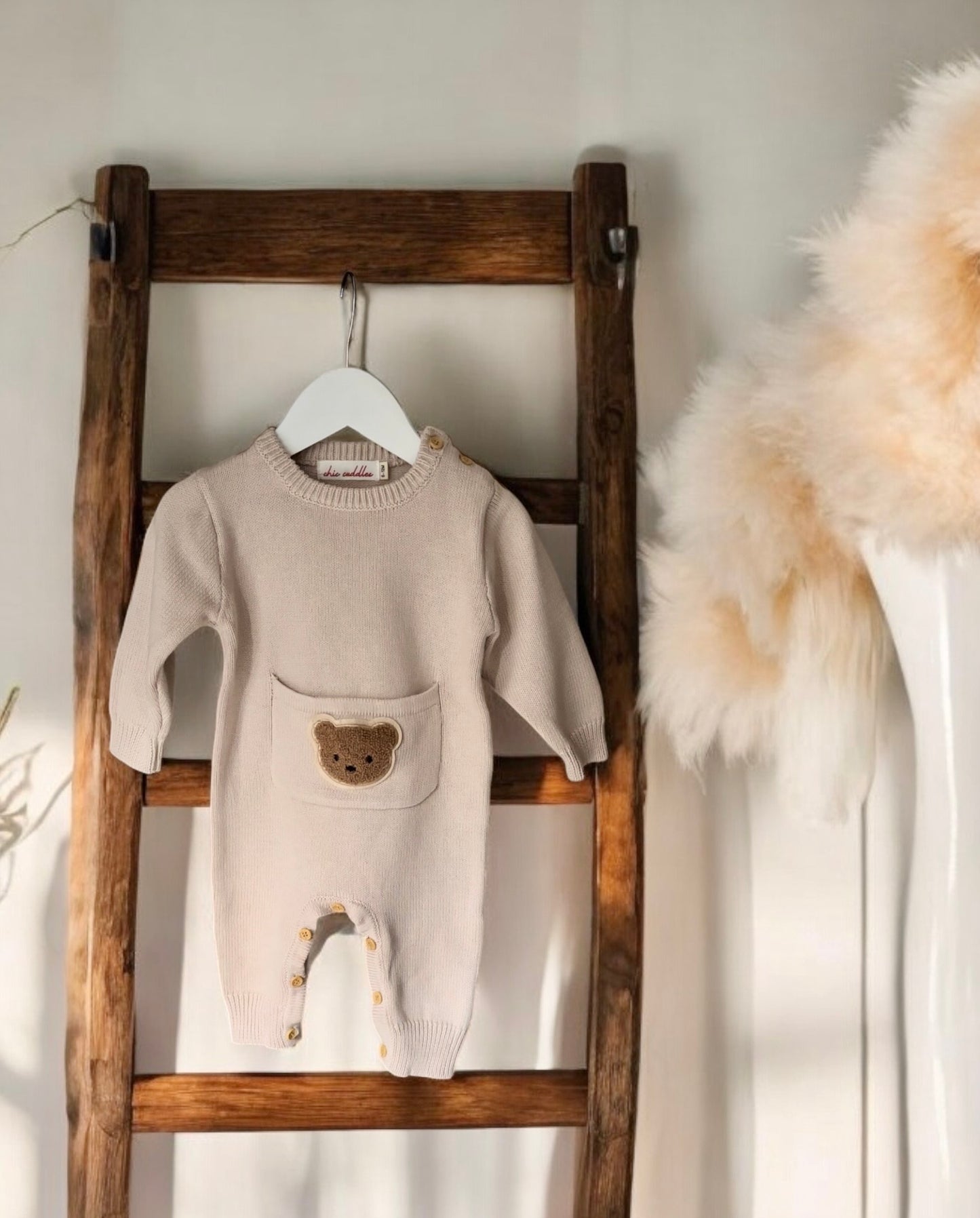 Beige Cosi Bear Pocket Onesie hanging on a wooden ladder, showing the full design