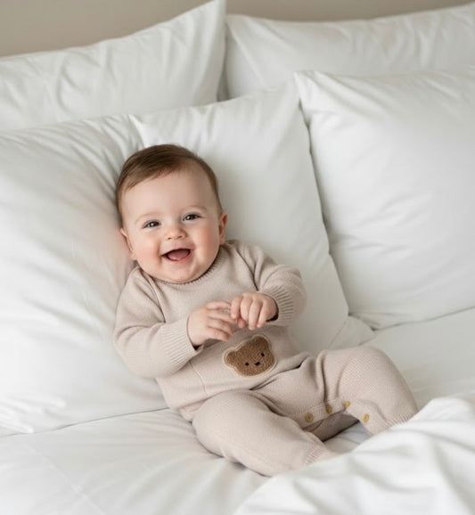 Smiling baby lying on a bed wearing the beige Cosi Bear Pocket Onesie with teddy pocket detail