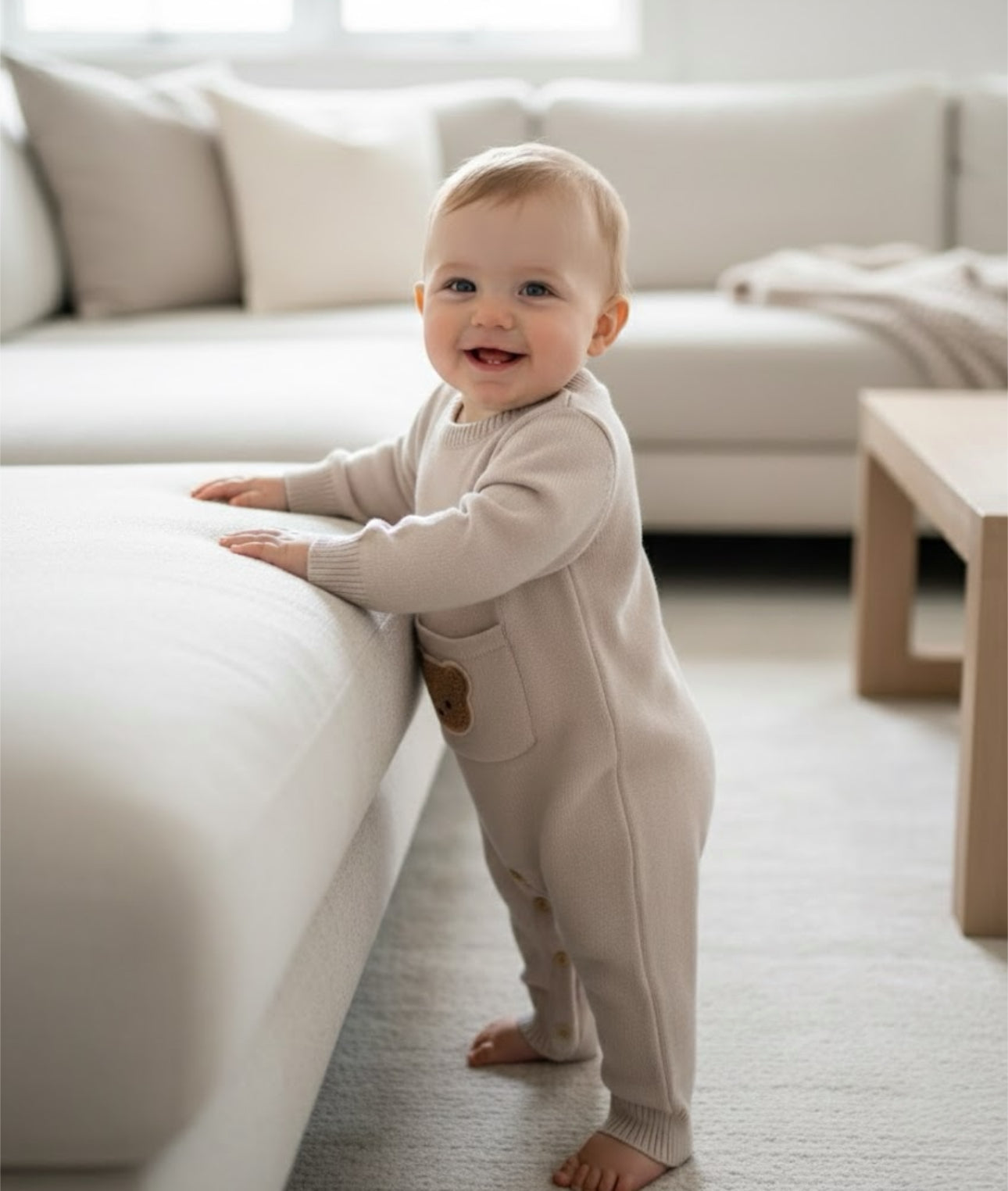 Baby standing and holding furniture while wearing the beige Cosi Bear Pocket Onesie